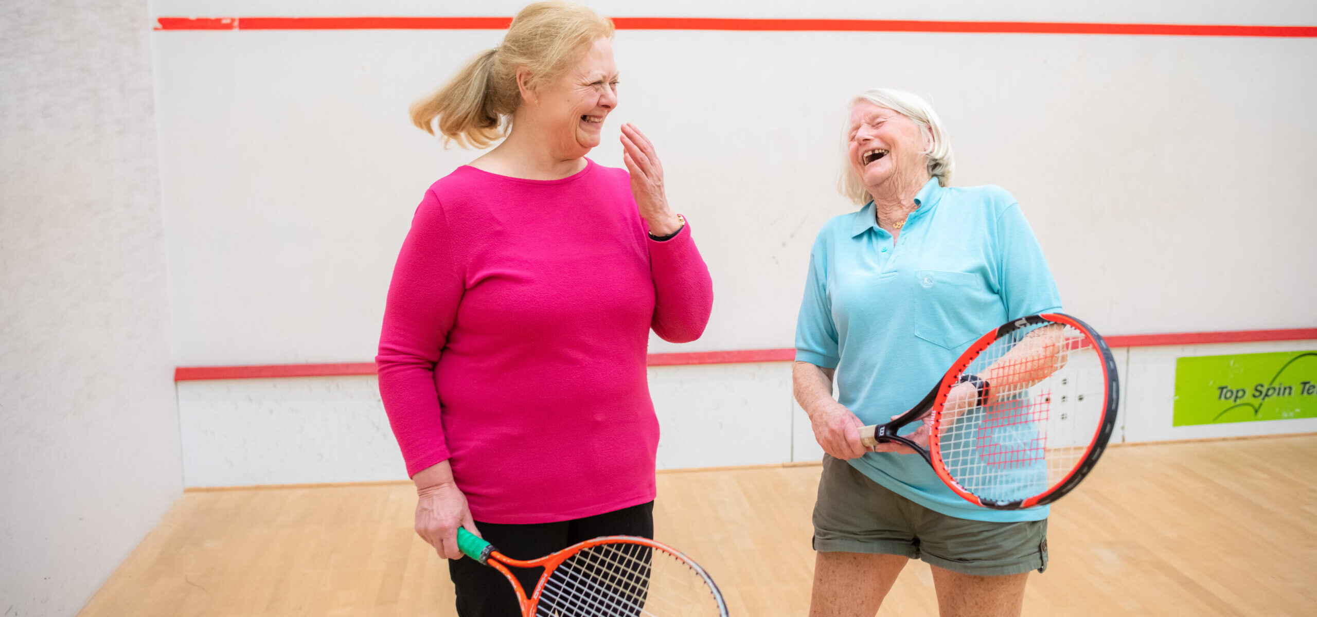 Two ladies on a squash court, laughing and holding tennis rackets.