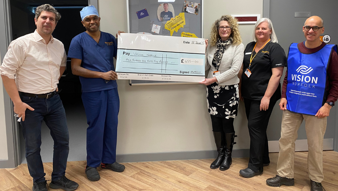 Five people smiling at the camera, displaying a large cheque made payable to Vision Norfolk for four hundred and fifty-five pounds, signed by Newmedica. The cheque is held by a man wearing surgical clothing and a lady with a Newmedica lanyard. One of the other men wears a Vision Norfolk tabard.