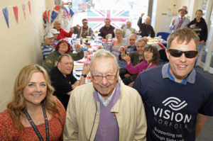 A representative from Blind Veterans UK (left) and Vision Norfolk (right) standing next to veteran and guest speaker Tony Wilkin (centre) inside the Vision Norfolk Great Yarmouth Hub. In the background Vision Norfolk service users, volunteers and staff are sitting at patriotically decorated tables raising their glasses to the camera.
