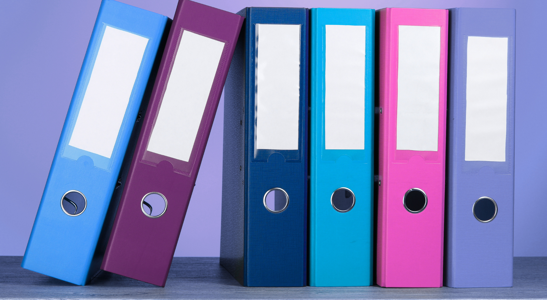 A stylised shot of six different coloured binders standing together side by side. The two binders on the left are leaning gently against the other four.