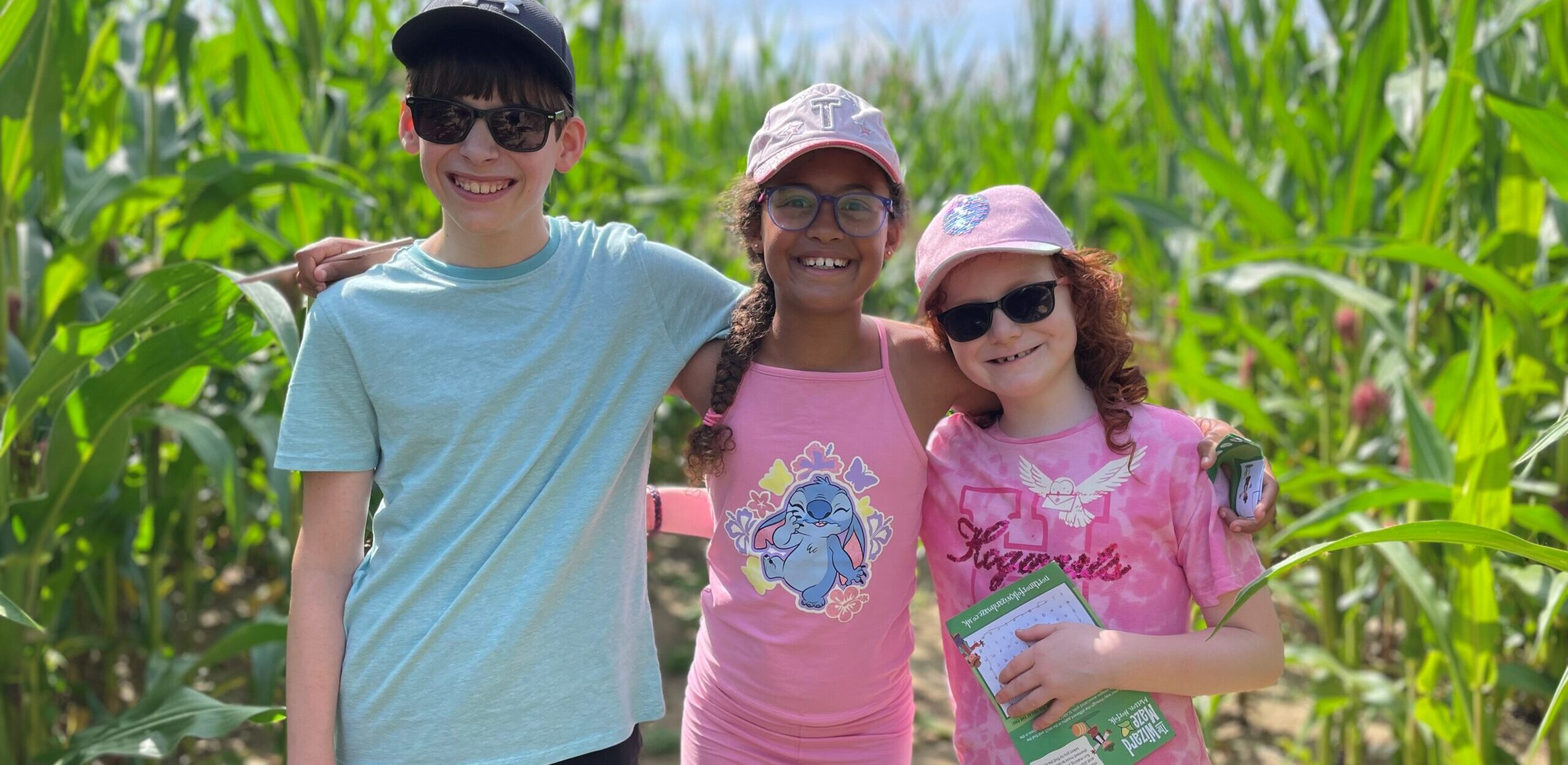 Three children stand side by side in a field surrounded by tall green plants in the sunshine. They are all wearing caps and summer clothes.
