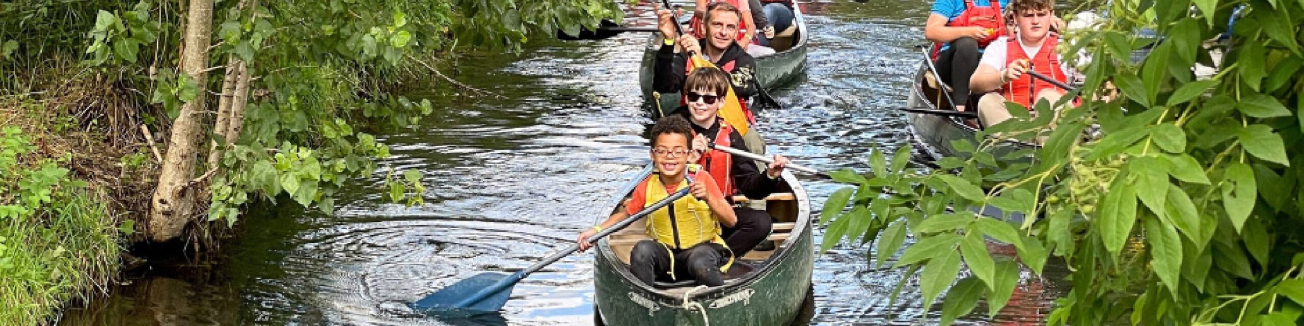 Several happy young children, rowing canoes along a river with adults supervising.