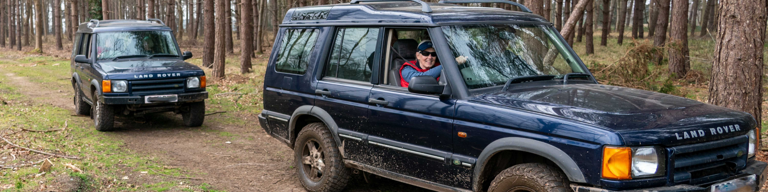 A woman in a red top smiling as she drives a four wheel Jeep. Another Jeep is following behind.