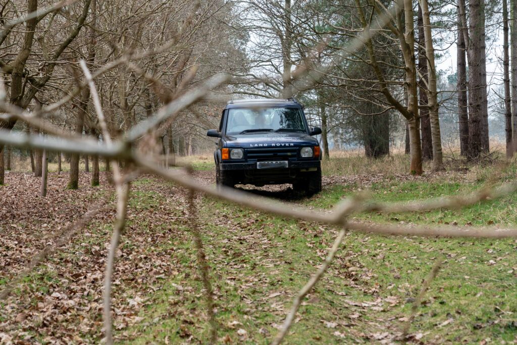 A 4x4 Land Rover vehicle driving through a wooded area viewed through branches in the foreground.