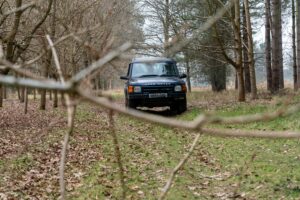 A 4x4 Land Rover vehicle driving through a wooded area viewed through branches in the foreground.