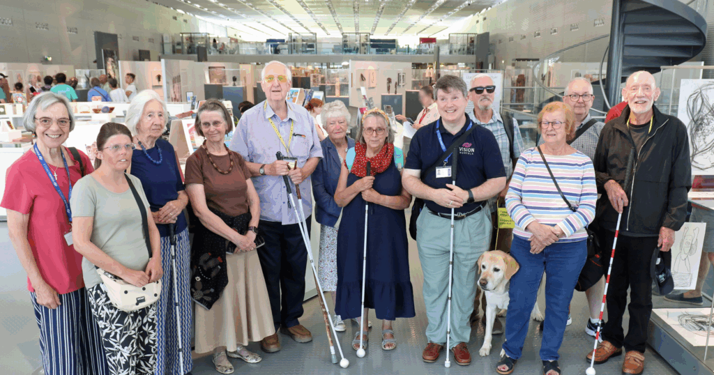 A group of 12 people, and one guide dog, standing inside the main exhibition room of the Sainsbury Centre in Norwich.