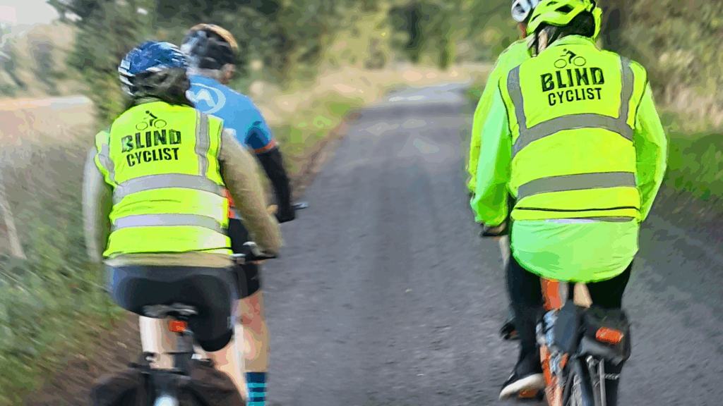 Two pairs of tandem cyclists riding on a road through trees. Both the cyclists at the back have reflective vests that say “Blind Cyclist”.