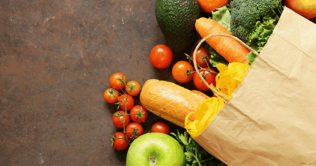 A brown paper grocery spilling over with food including pasta, bread, tomatoes, apples, broccoli, carrots and an avocado