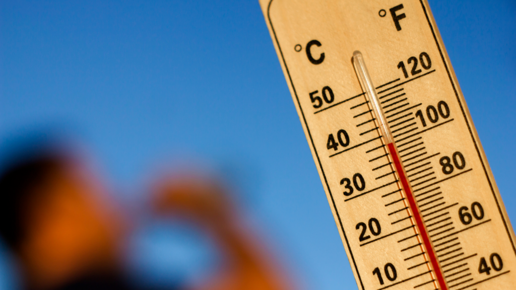 A wooden thermometer showing a very high temperature, around 37 degrees Celsius. In the background there is a blurry outline of someone drinking water.