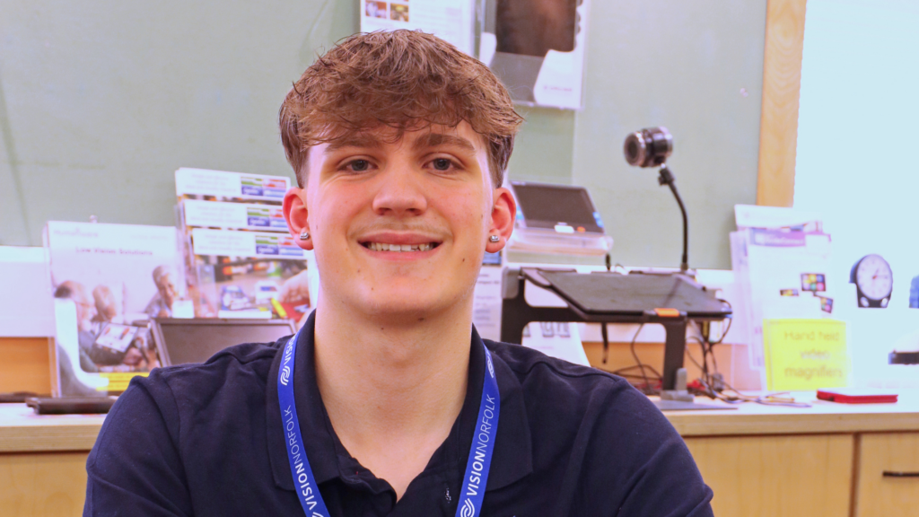 Volunteer Max Bevan siting in the Norwich Equipment Centre with pamphlets and pieces of low vision equipment behind him.