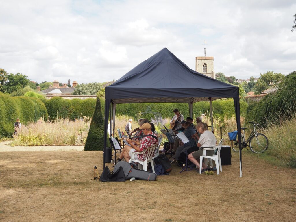 TATS Buskers playing music under a gazebo on the main lawn. The herb garden and some hedges are behind them.