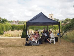 TATS Buskers playing music under a gazebo on the main lawn. The herb garden and some hedges are behind them.