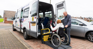 A woman in a wheelchair using the lift to get on to a van from Norwich Door to Door. A man is standing to her right assisting her.