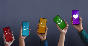 Hands holding up smartphones with a scale of emotions on the screens, from a red frowning face to a green smiling face. On the far right is a phone with a heart on it.